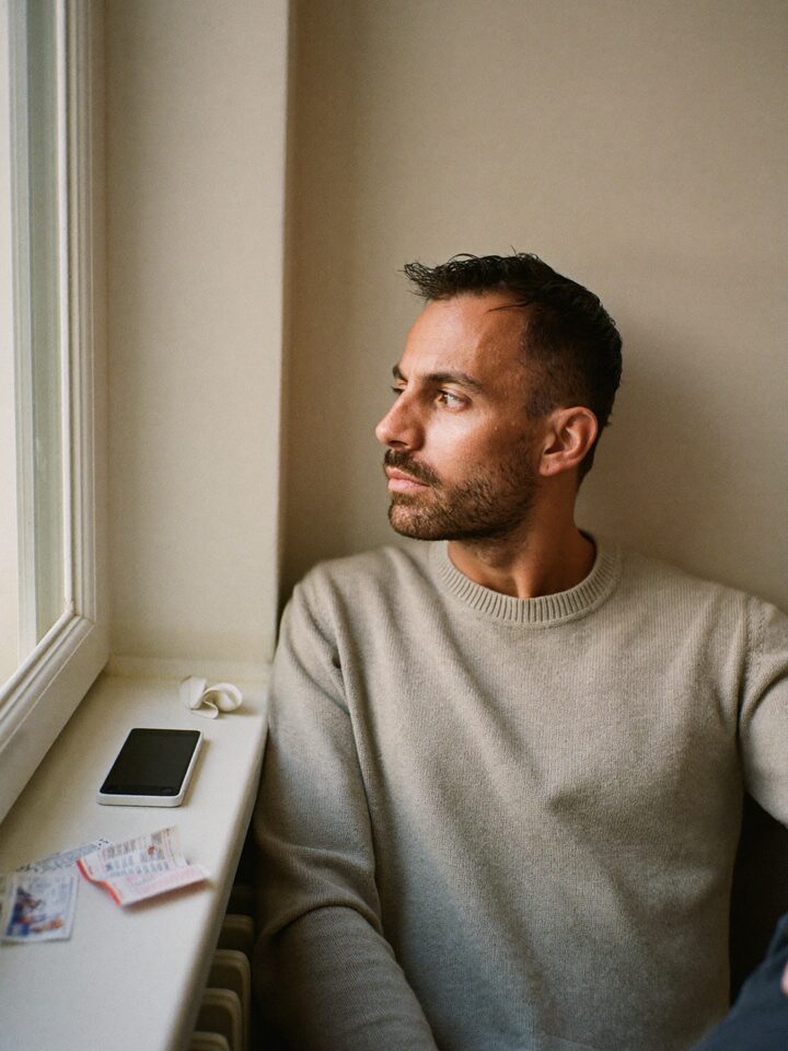 Ein Mann sitzt an einem Fenster und blickt nachdenklich nach draußen; neben ihm liegen sein abgelegtes Smartphone und mehrere Wett- und Lottoscheine.